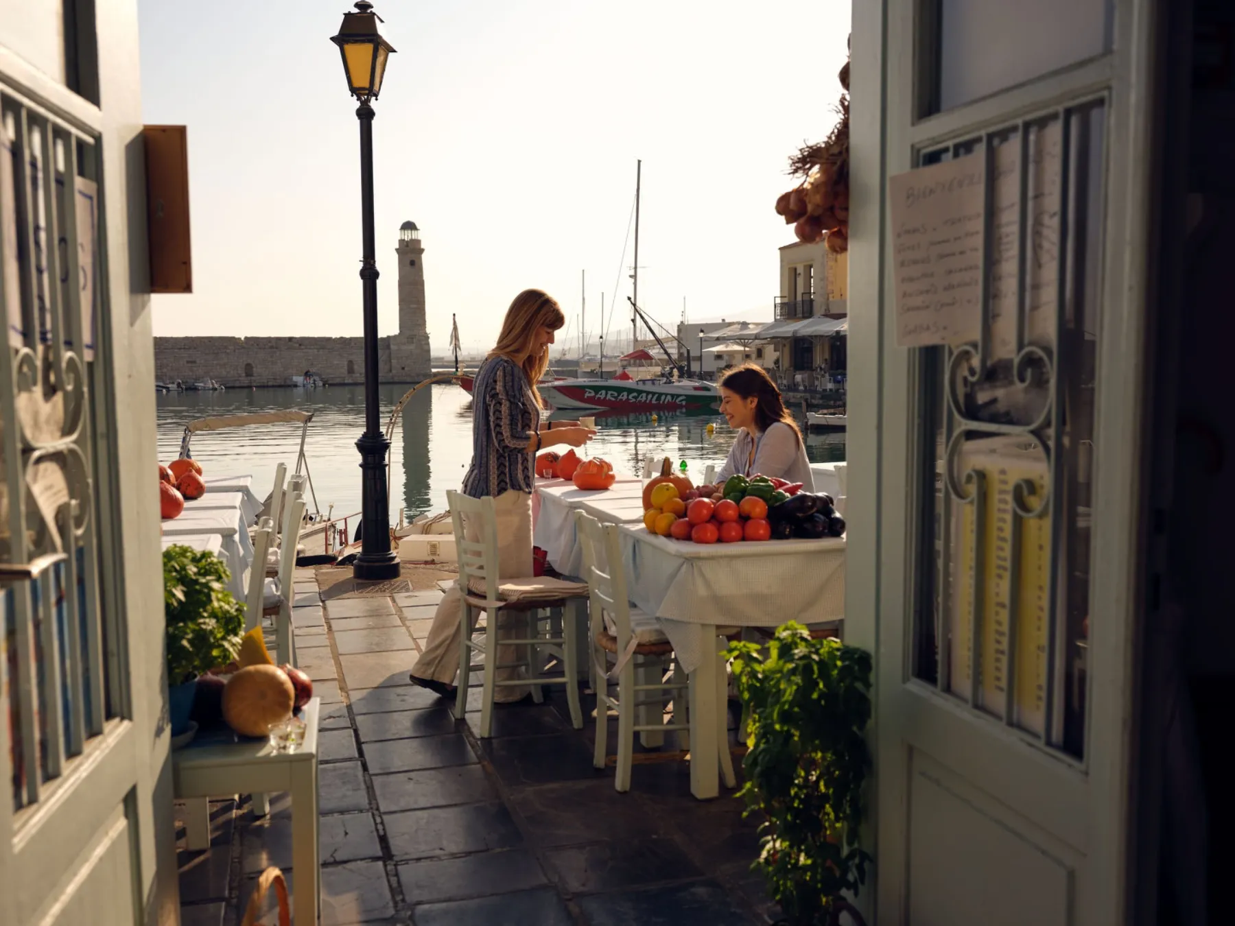 Two women at a harbourside taverna table in Rethymno — the Venetian lighthouse and fishing boats behind them, ripe tomatoes and vegetables on the cloth