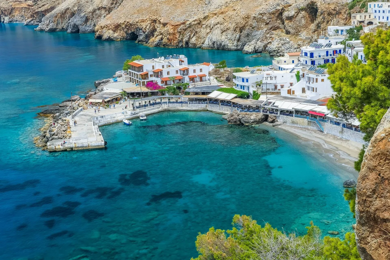 Chora Sfakion from above: the small harbour, white houses and waterfront tavernas, and a sandy cove in clear turquoise water under the cliffs