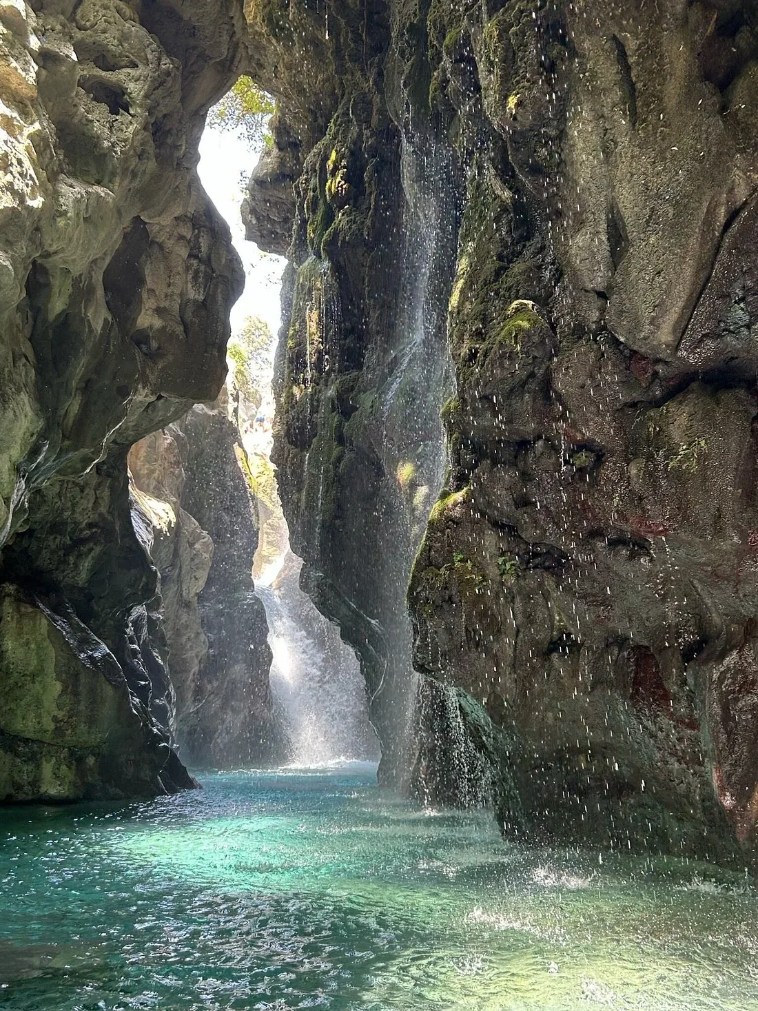 A waterfall pouring down the rock walls into the green pool of Kourtaliotiko Gorge